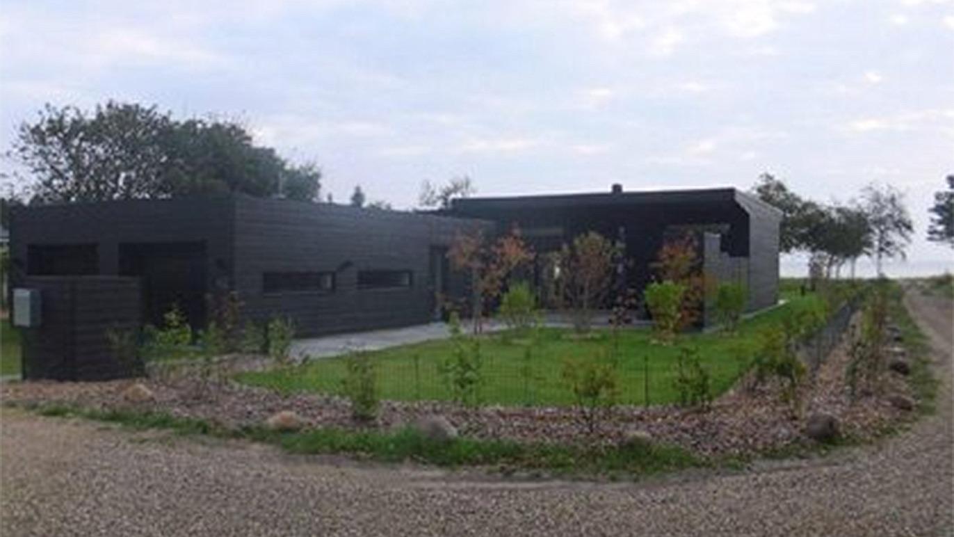 Moderne sort hus med have og grusvej, omgivet af træer og buske under en skyet himmel.