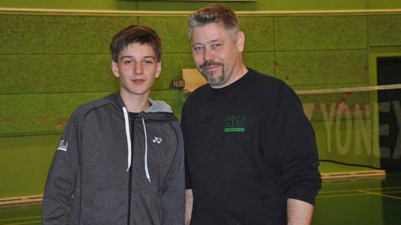 To personer står sammen i en badmintonhal, en ung dreng i grå hættetrøje og en mand med skæg i sort trøje.