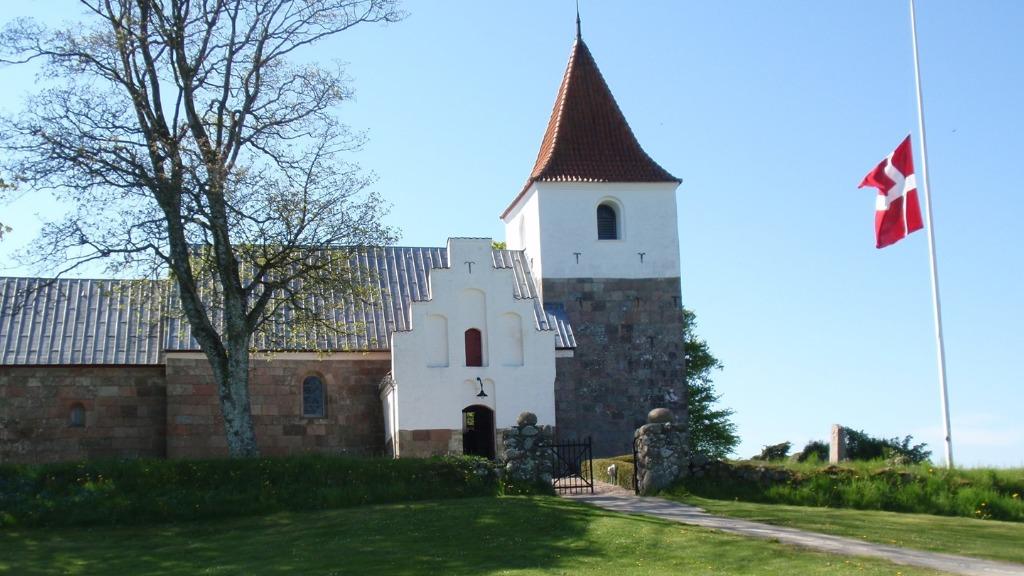En historisk kirke med en spids tårn og dansk flag, omgivet af grønne områder og træer.