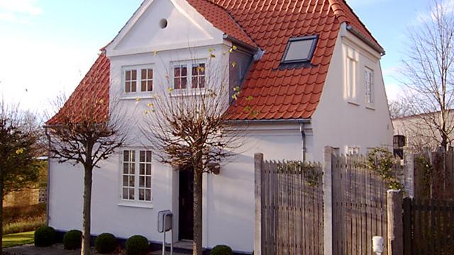 Et hvidt hus med rød tag og træhegn, omgivet af små træer og en klar blå himmel.