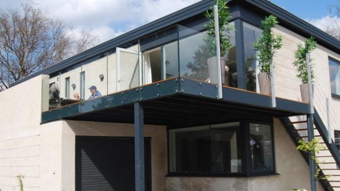 Moderne hus med glasfacade, balkon og grønne planter, beliggende i et roligt område under en blå himmel.