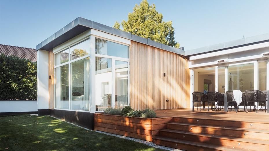 Moderne hus med træfacade og store vinduer, beliggende i en have med terrasse og udendørs siddepladser.