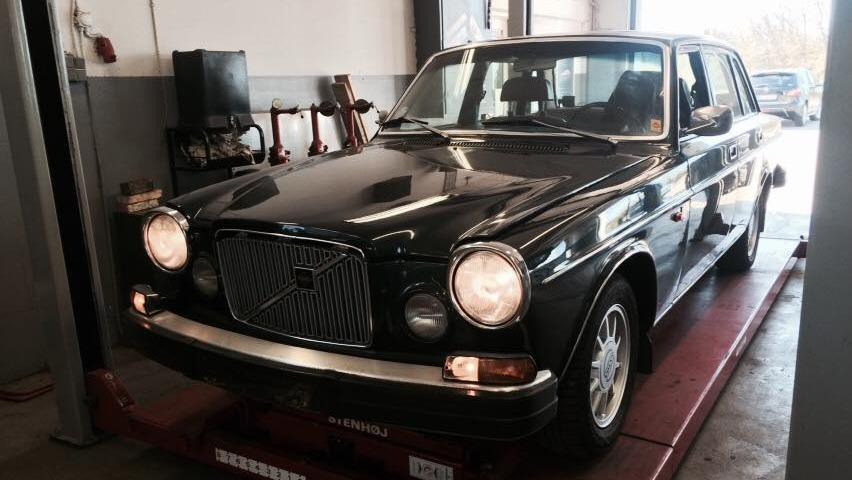 En sort Volvo bil står på en lift i et værksted, med lysene tændt og en klassisk design.