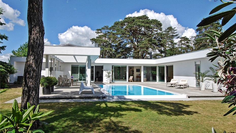 Moderne hus med swimmingpool og grøn have omgivet af træer under en blå himmel.