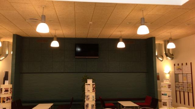 Indendørs café med grøn væg, lamper i loftet og et tv på væggen. Bord og stole i moderne design.