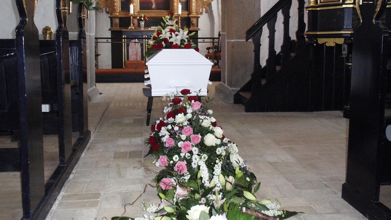 Kiste med blomsterarrangement i kirke, omgivet af bænke og alter, skaber en stille og respektfuld atmosfære.