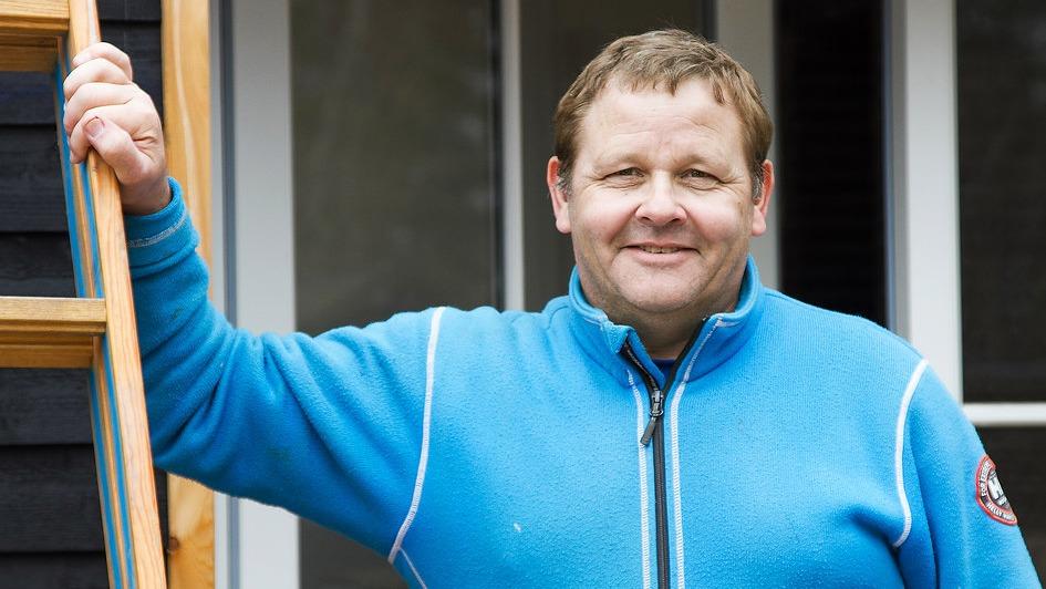 En smilende mand i en blå trøje står ved en stige foran et hus.