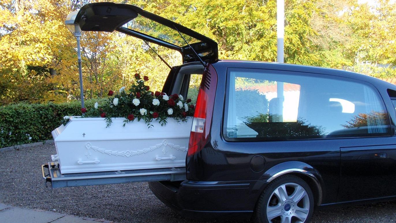 En sort ligvogn med en hvid kiste fyldt med blomster, der står åben i en smuk efterårsindstilling.