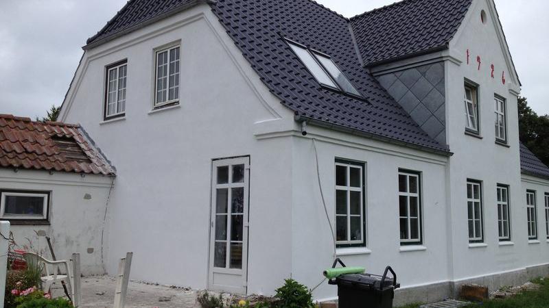 Hvidt hus med sort tag og vinduer, bygget i 1722, beliggende i en have med møbler og affaldscontainer.