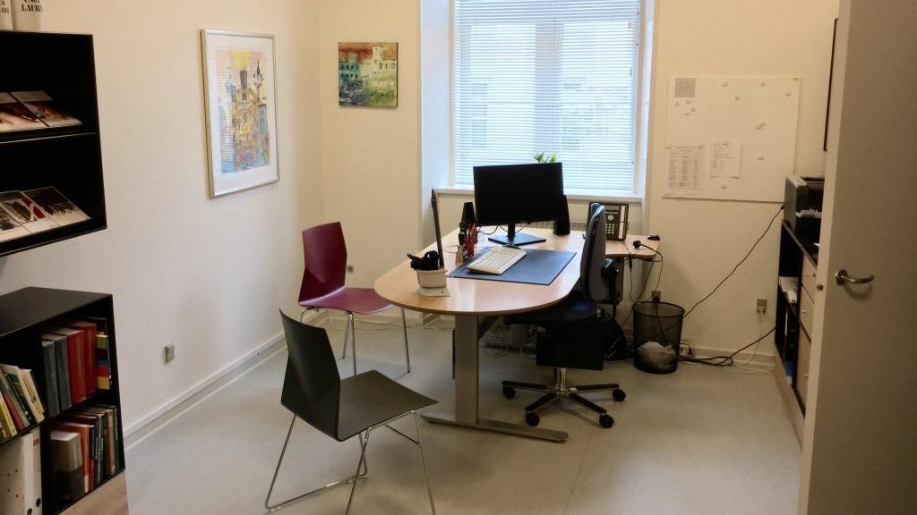 Et kontor med et skrivebord, en computer, to stole og reoler fyldt med bøger. Vindue med naturligt lys.