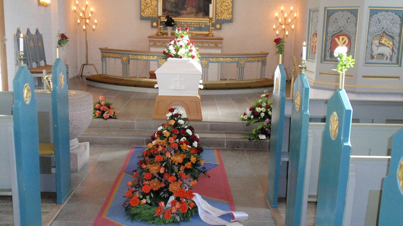 Begravelsesceremoni i kirke med blomsterdekorationer og kiste i midten af rummet.