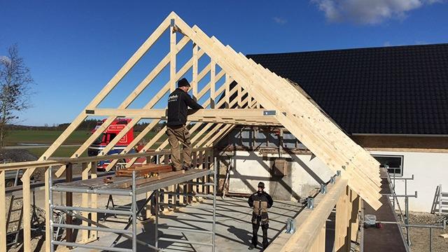To mænd arbejder på at bygge et tag med trærammer på en nybygget hus. Scenen viser stilladser og en blå himmel.