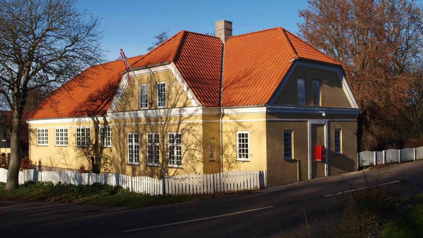 Et gult hus med rødt tag og hvidt hegn, beliggende ved vejen under en klar blå himmel.