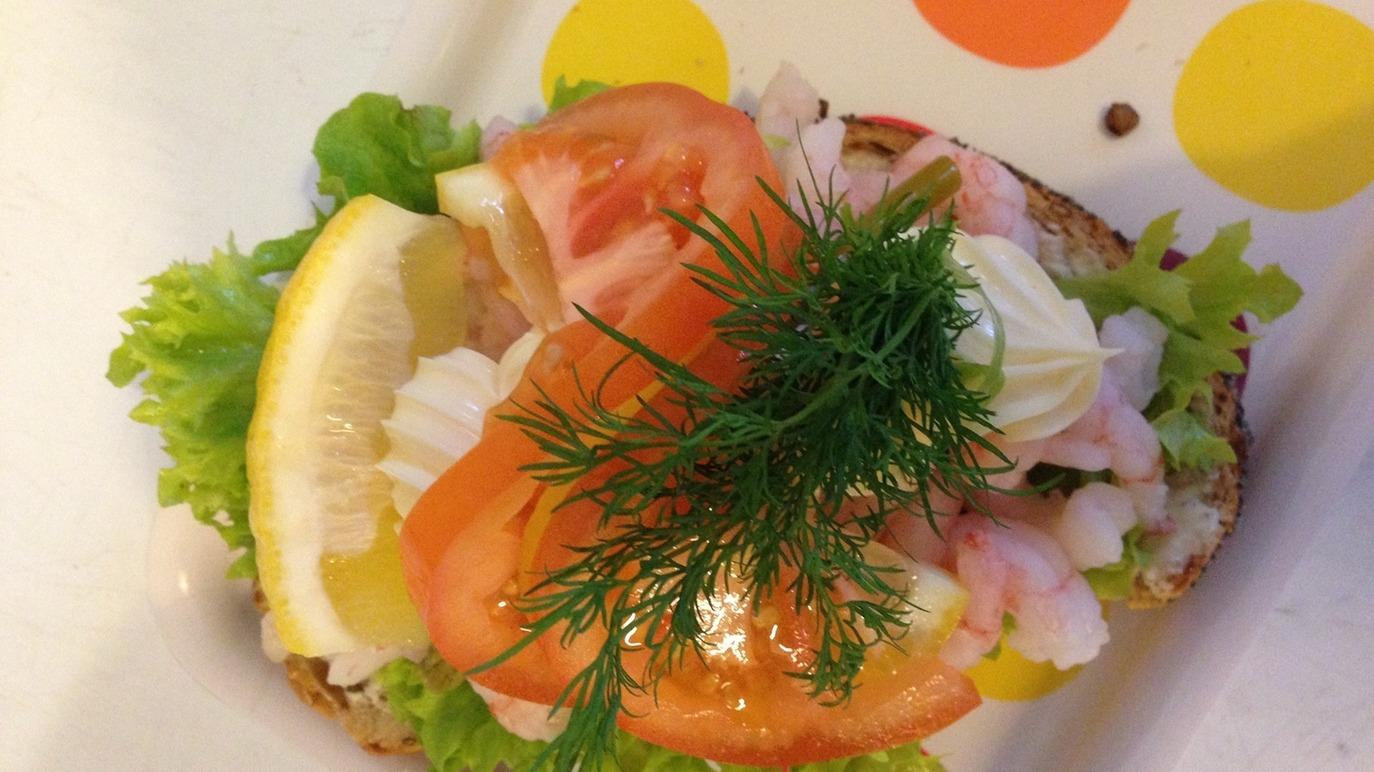 En smørrebrød med rejer, salat, tomat, citron og dild på en farverig tallerken.