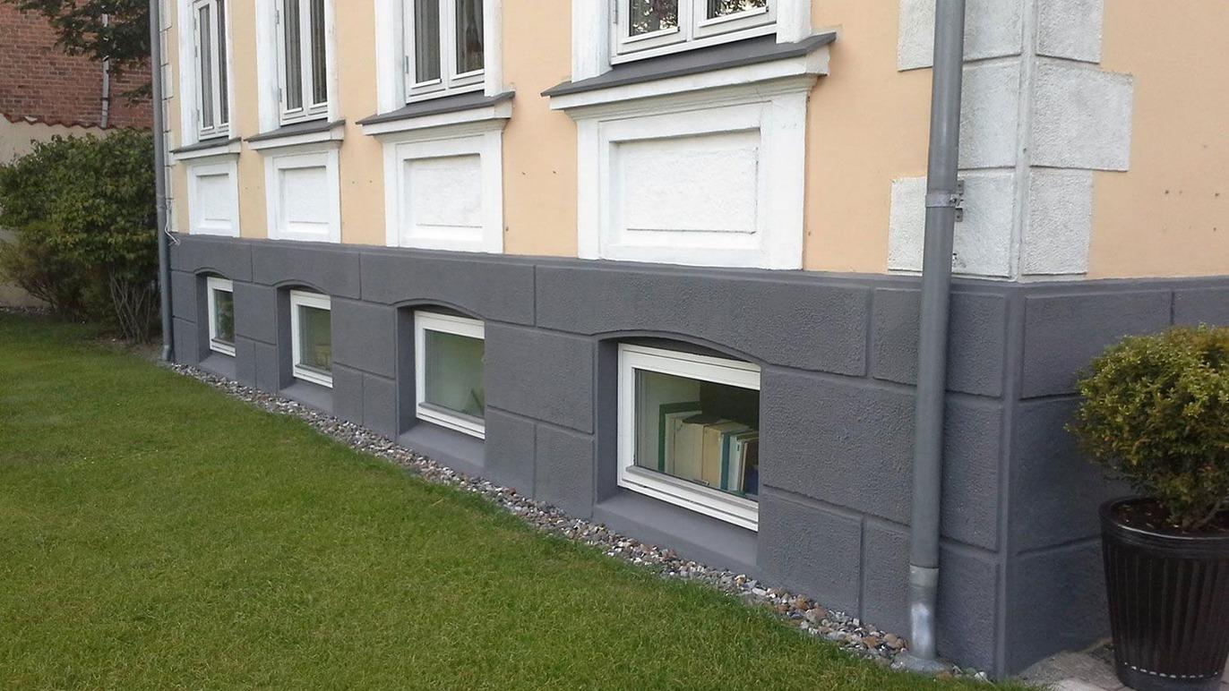 Facade af bygning med grå sokkel og vinduer, der er delvist under jorden, omgivet af græs og småsten.