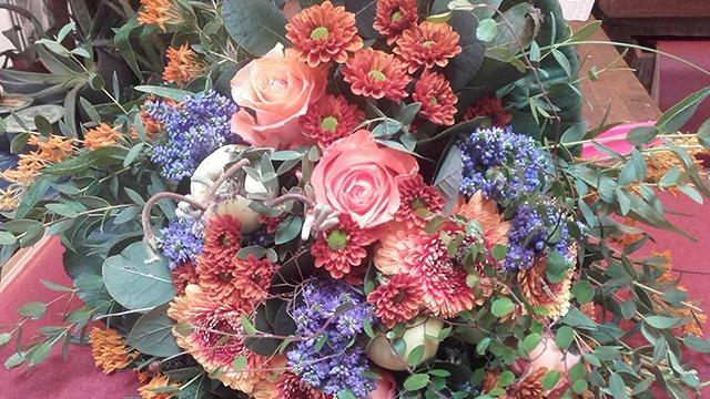 En farverig buket med roser, chrysantemum og blå blomster, omkranset af grønne blade.