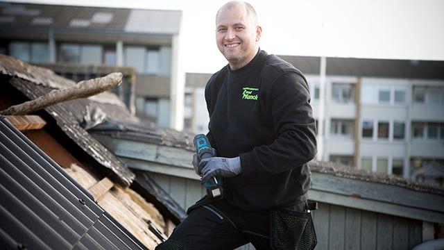 Håndværker arbejder på taget med en boremaskine, iført sort trøje og handsker, med bygninger i baggrunden.