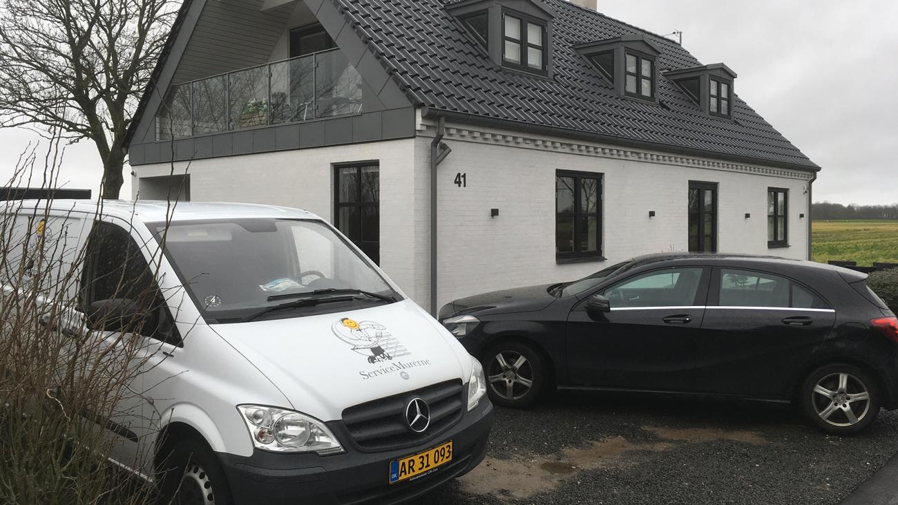 Moderne hus med sort tag, en hvid varevogn og en sort bil parkeret foran. Grønt landskab i baggrunden.