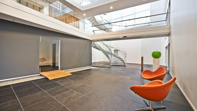 Moderne foyer med glasloft, trappe, sorte fliser og orange stole.