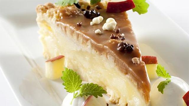 Lækkert stykke kage med karamel, chokolade og frugt, serveret med flødeskum og mynte på en hvid tallerken.