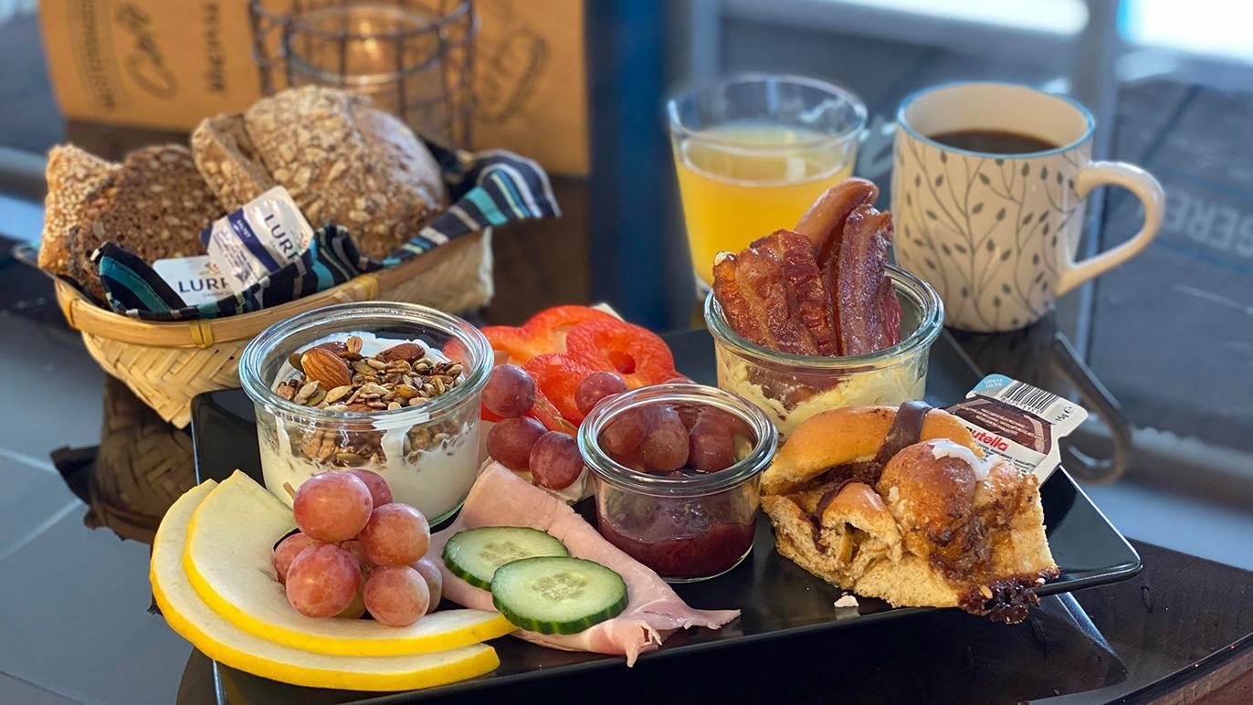 En lækker morgenmadstallerken med yoghurt, granola, frugt, brød, pålæg og drikkevarer.