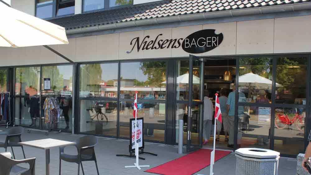 Indgangen til Nielsens Bageri med udendørs siddepladser og danske flag foran.