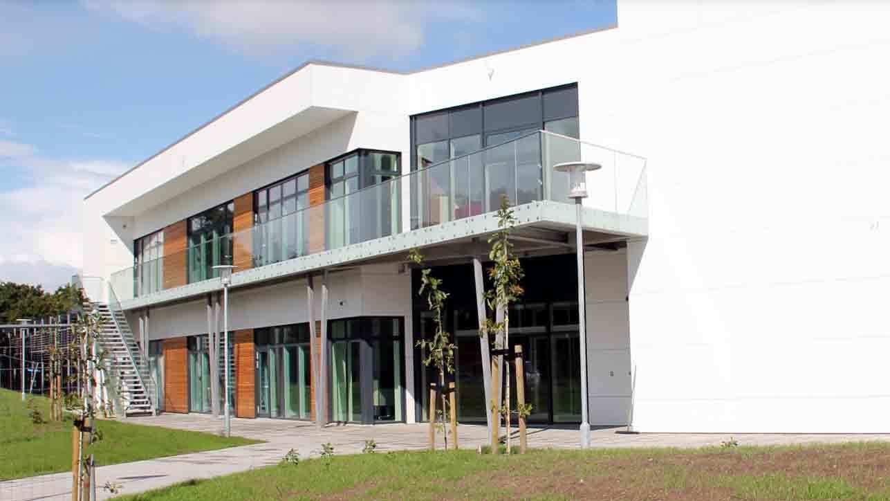Moderne bygning med glasfacade og trædetaljer, omgivet af grøn græsplæne og små træer.
