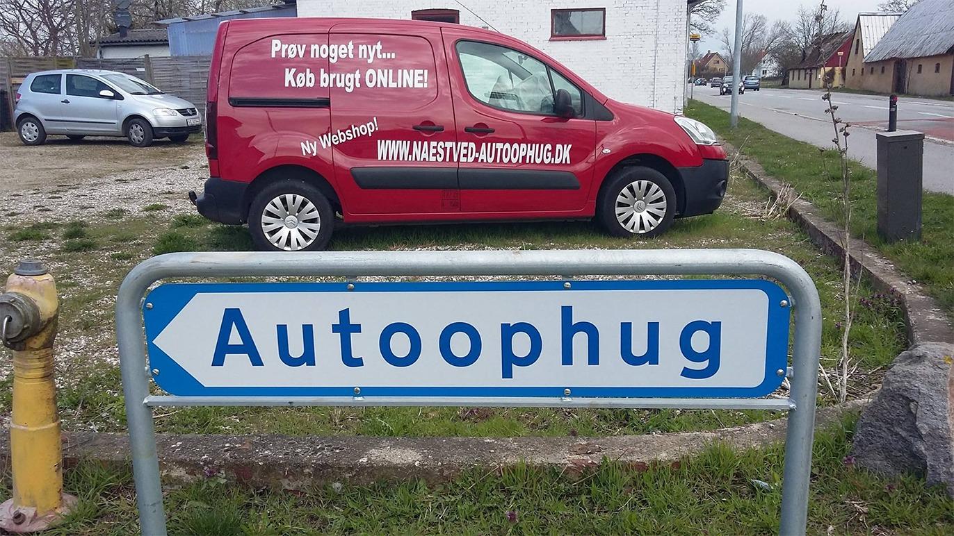 Rød varevogn med tekst om autohug står ved skilt med teksten "Autohug" i en landsby.