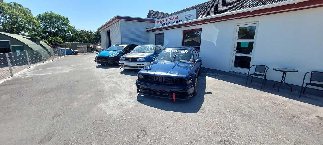 En sort bil parkeret foran en bilforhandler med flere biler i baggrunden og en solrig dag.