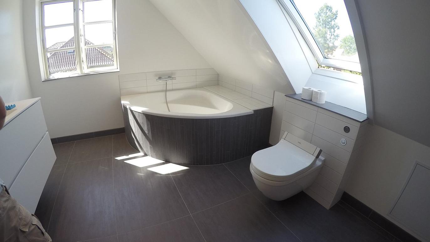 Lyst badeværelse med skrå vægge, badekar og moderne toilet ved vinduet.