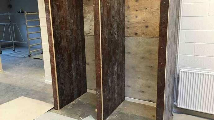 Toiletvægge i træ med rå overflader, der er delvist opført i et rum med grå gulvbelægning.