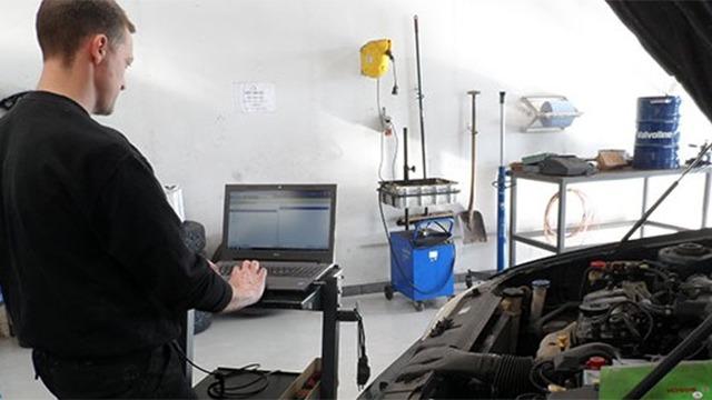 En mekaniker arbejder på en computer ved en bil i et værksted med værktøj og udstyr i baggrunden.