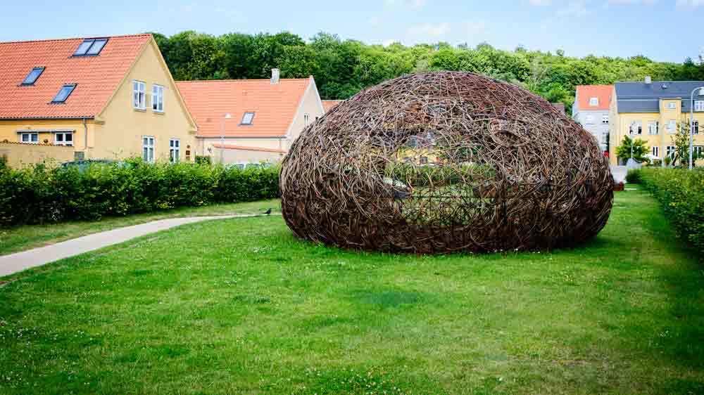 En stor, rund rede lavet af grene står på en græsplæne mellem huse og træer.