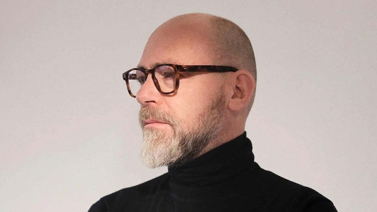 Mand med briller og skæg iført sort turtleneck, står i profil mod en lys baggrund.