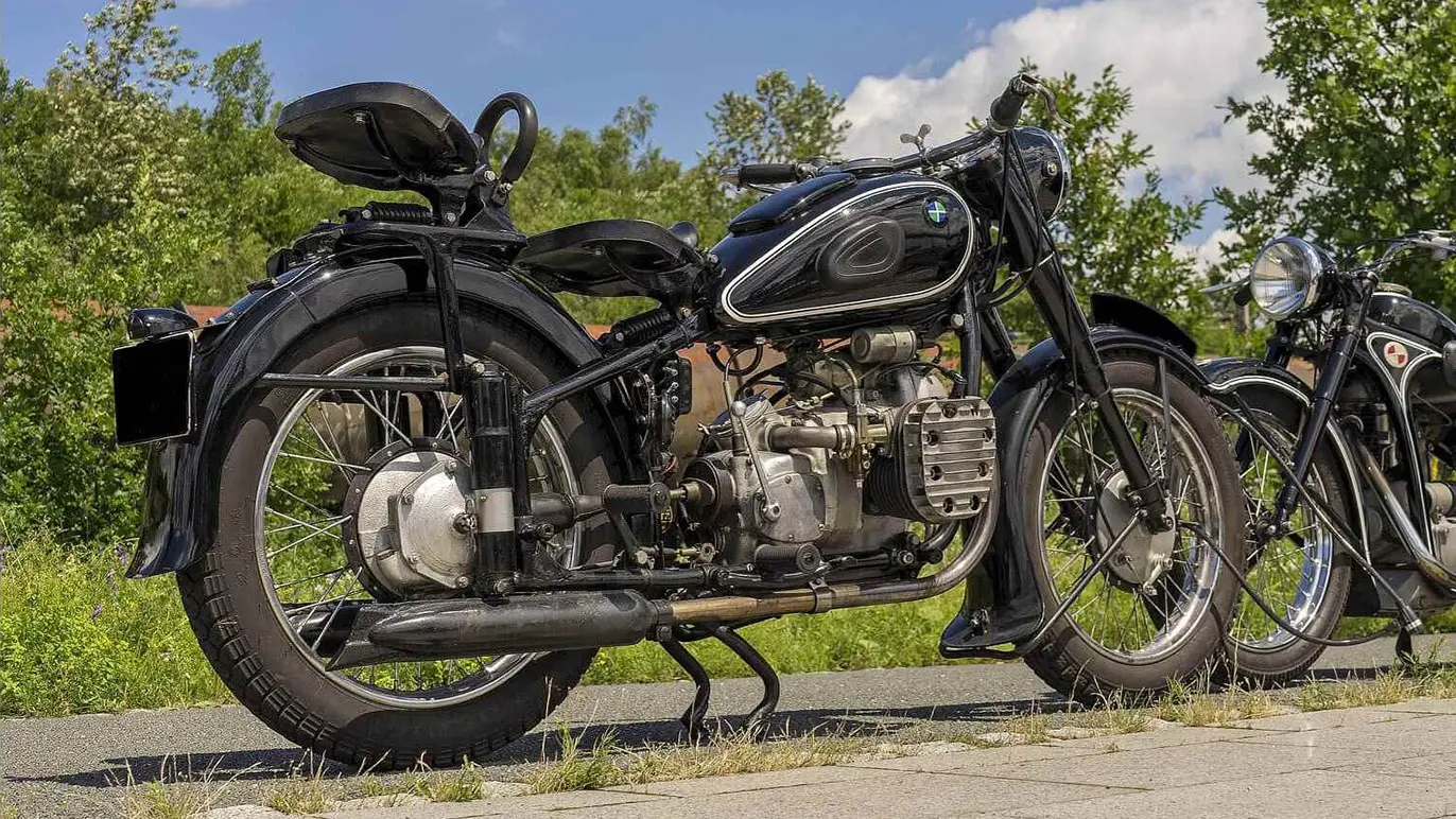 En klassisk motorcykel parkeret langs en vej med grønne træer i baggrunden.
