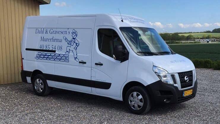 Hvid varevogn med logo og tekst, parkeret ved en bygning på en grusvej med grønne marker i baggrunden.
