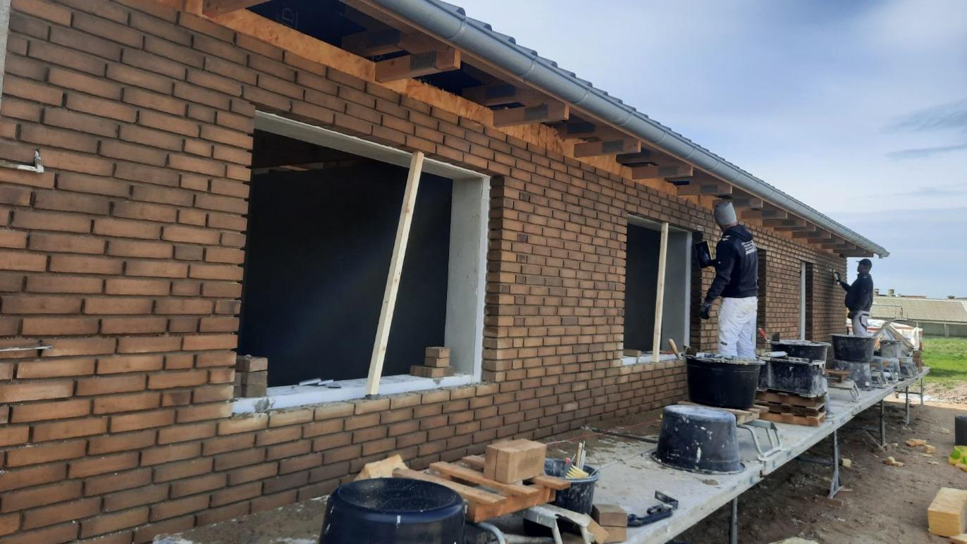 Byggeri af murstenshus med håndværkere, der arbejder på vinduer og facade, samt materialer på byggepladsen.