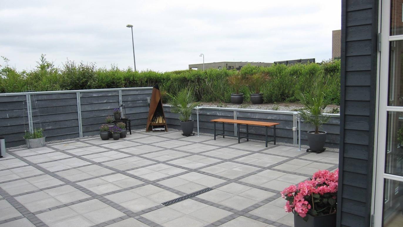 En terrasse med fliser, planter i krukker og en bænk, omgivet af grønne buske og et hegn.