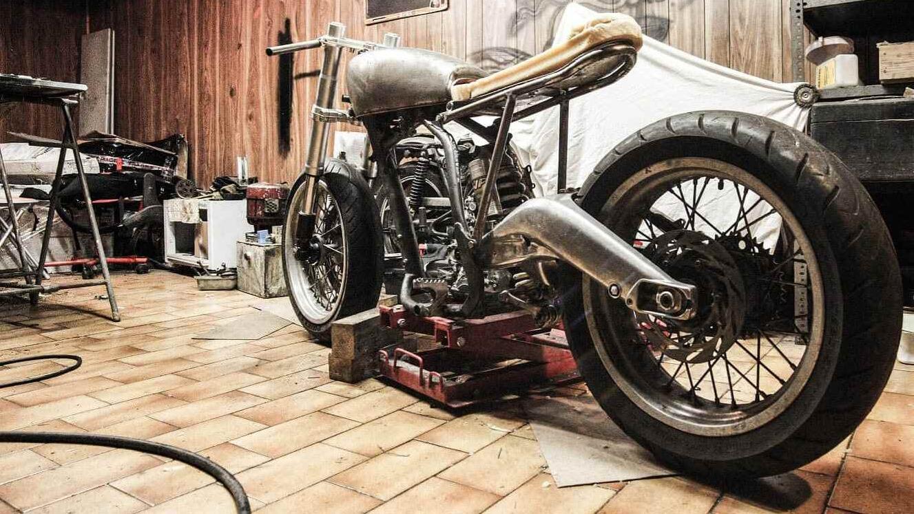 En motorcykel med metalramme og guld sæde står i en garage med flisegulv og værktøj i baggrunden.