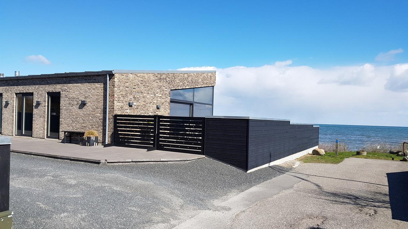 Moderne hus ved kysten med stenfacade og udsigt over havet under en blå himmel.