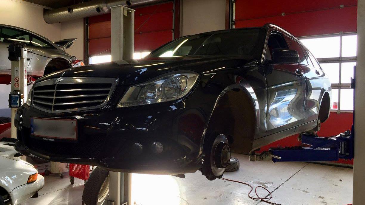 En sort Mercedes bil hævet på en lift i et autoværksted med værktøj og biler i baggrunden.