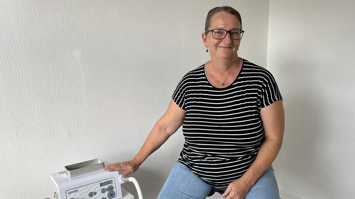 Kvinde med briller sidder ved en maskine i et lyst rum, iført en stribet t-shirt og jeans.