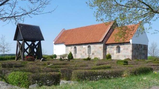 En kirke med rødt tag og stenfacade, omgivet af en velplejet hæk og en træklokketårn i baggrunden.
