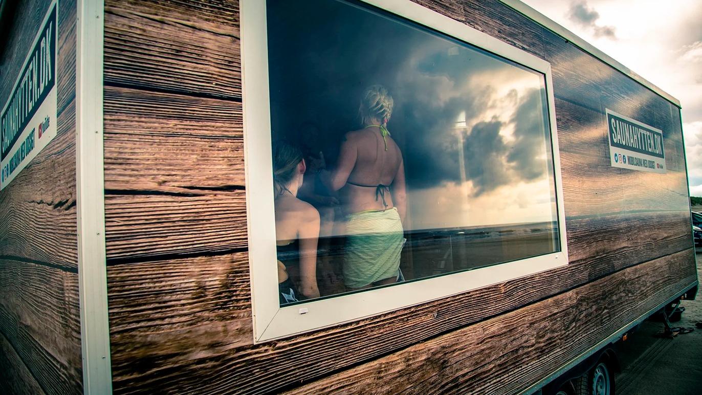 To personer kigger ud af et vindue i en træbeklædt trailer med en dramatisk himmel i baggrunden.