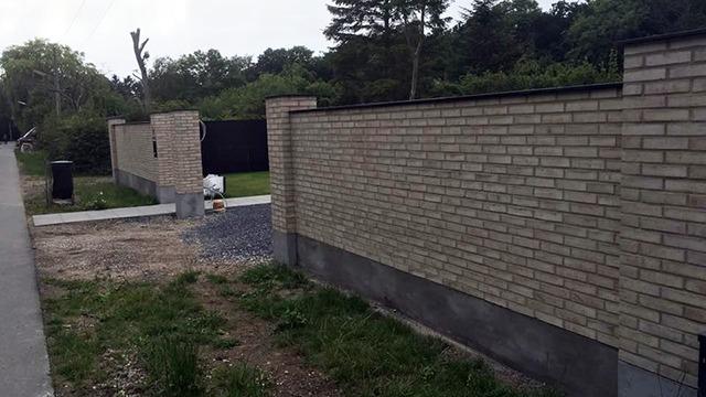 Mur af mursten med græsplæne og træer i baggrunden, beliggende ved en vej.