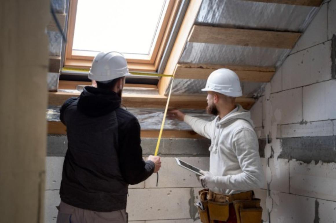 To personer i hjelme måler og arbejder på en loftsrenovering med isolering og vindue.