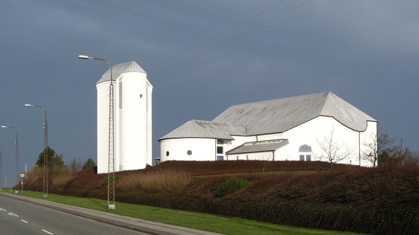 En moderne hvid kirke med et tårn, beliggende ved en vej under en mørk himmel.