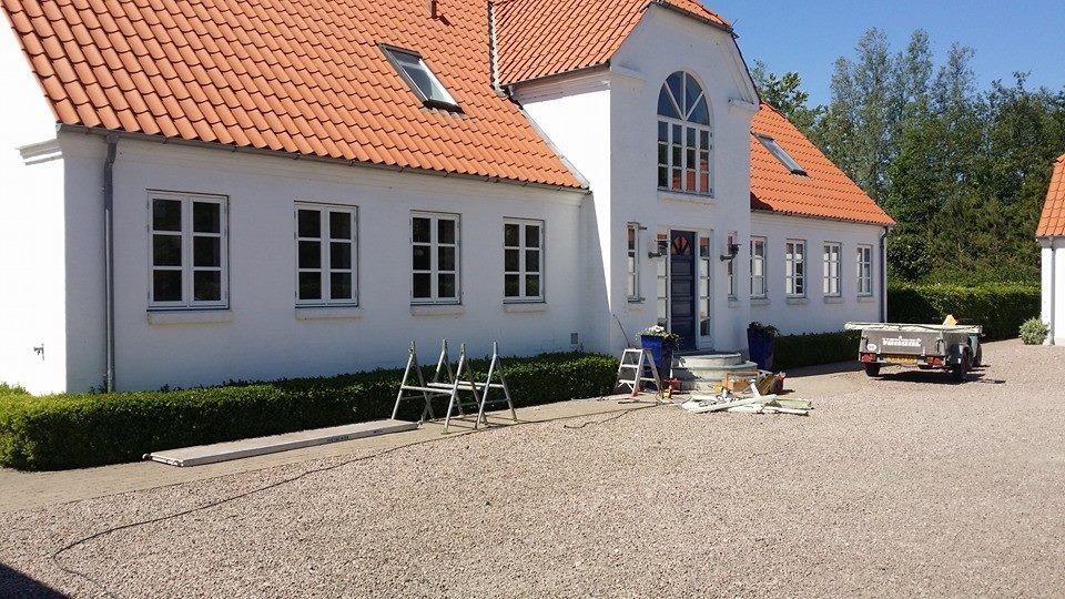 Hvidt hus med rød tag, havearbejde i gang og byggeudstyr på grusbelagt indkørsel.