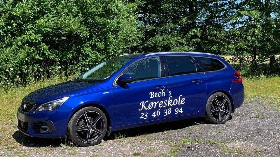 Blå bil med teksten "Bech's Køreskole" og telefonnummeret 23 46 38 94, parkeret i et grønt område.