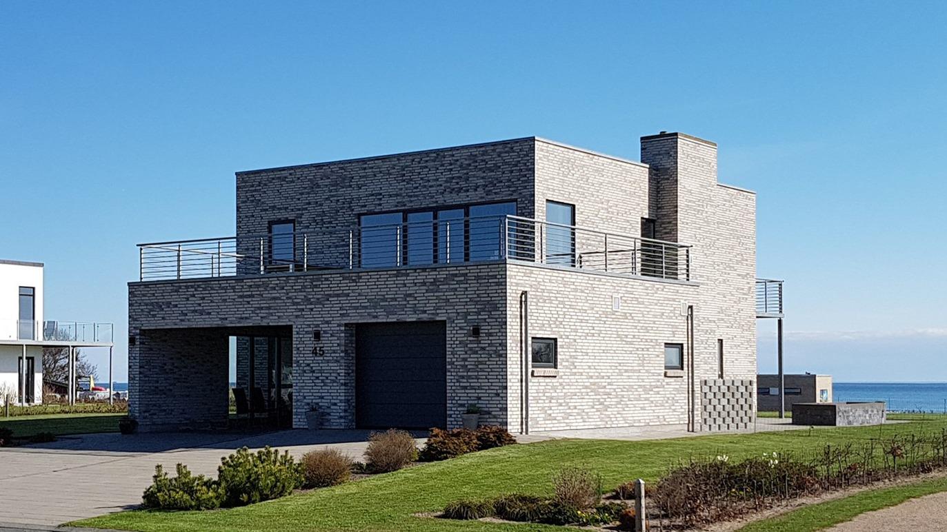 Moderne hus med grå mursten og store vinduer, beliggende ved kysten med havudsigt.